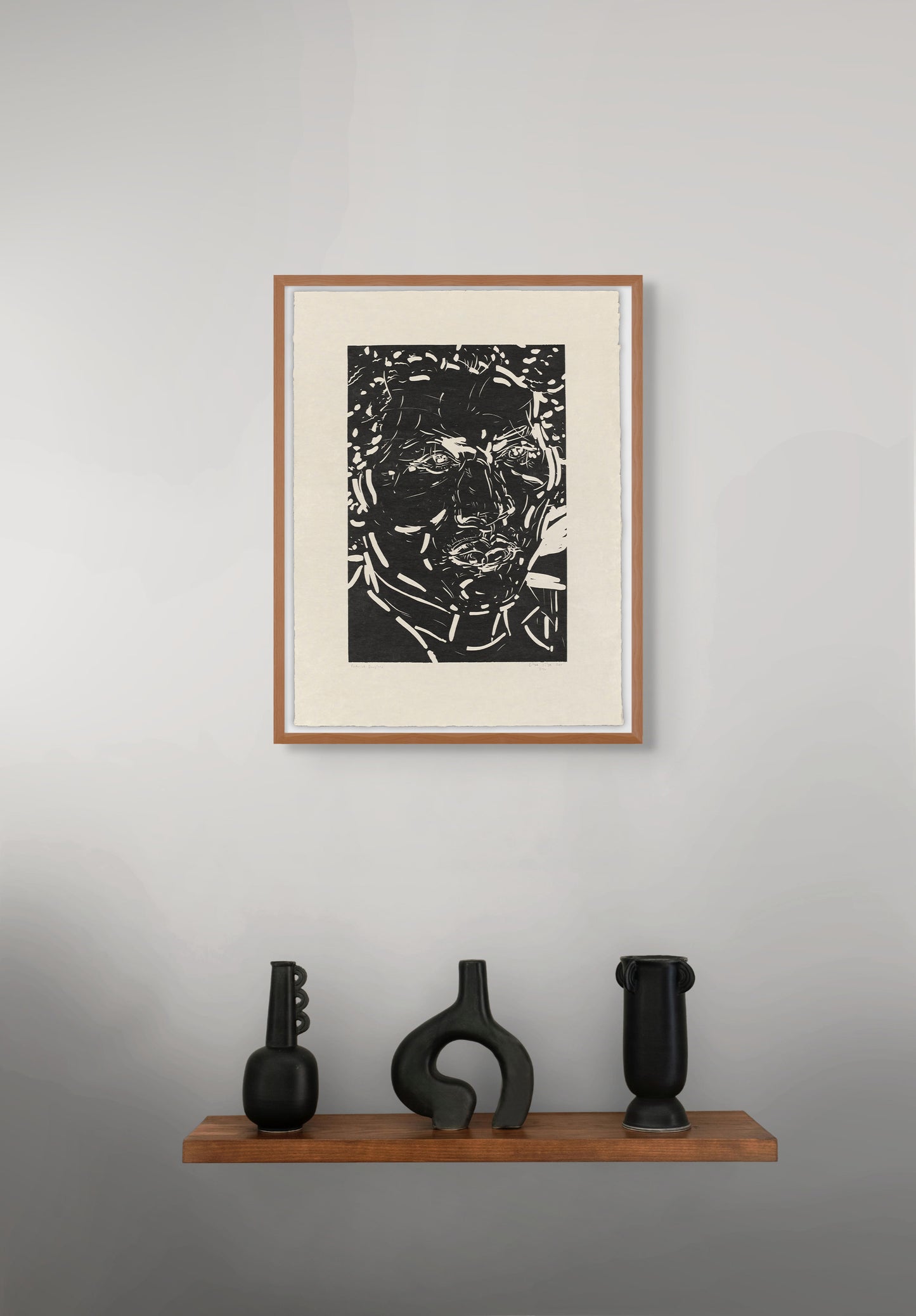 Framed black and white portrait by Elizabeth Peyton on a gray wall with three black sculptures on a wooden shelf below.