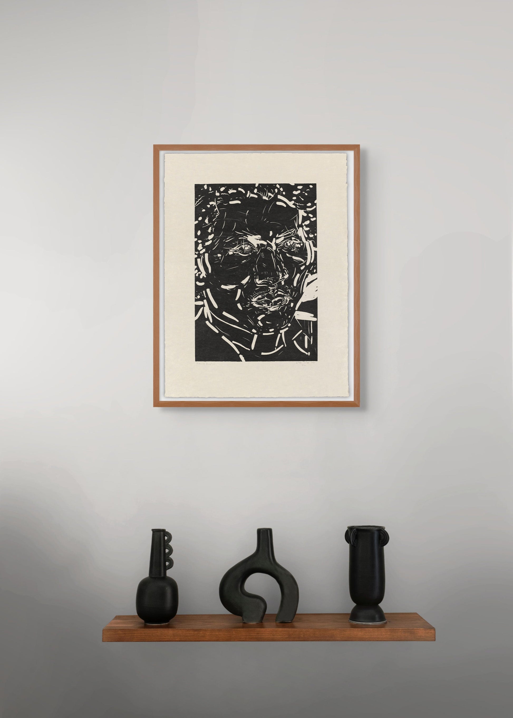 Framed black and white portrait by Elizabeth Peyton on a gray wall with three black sculptures on a wooden shelf below.