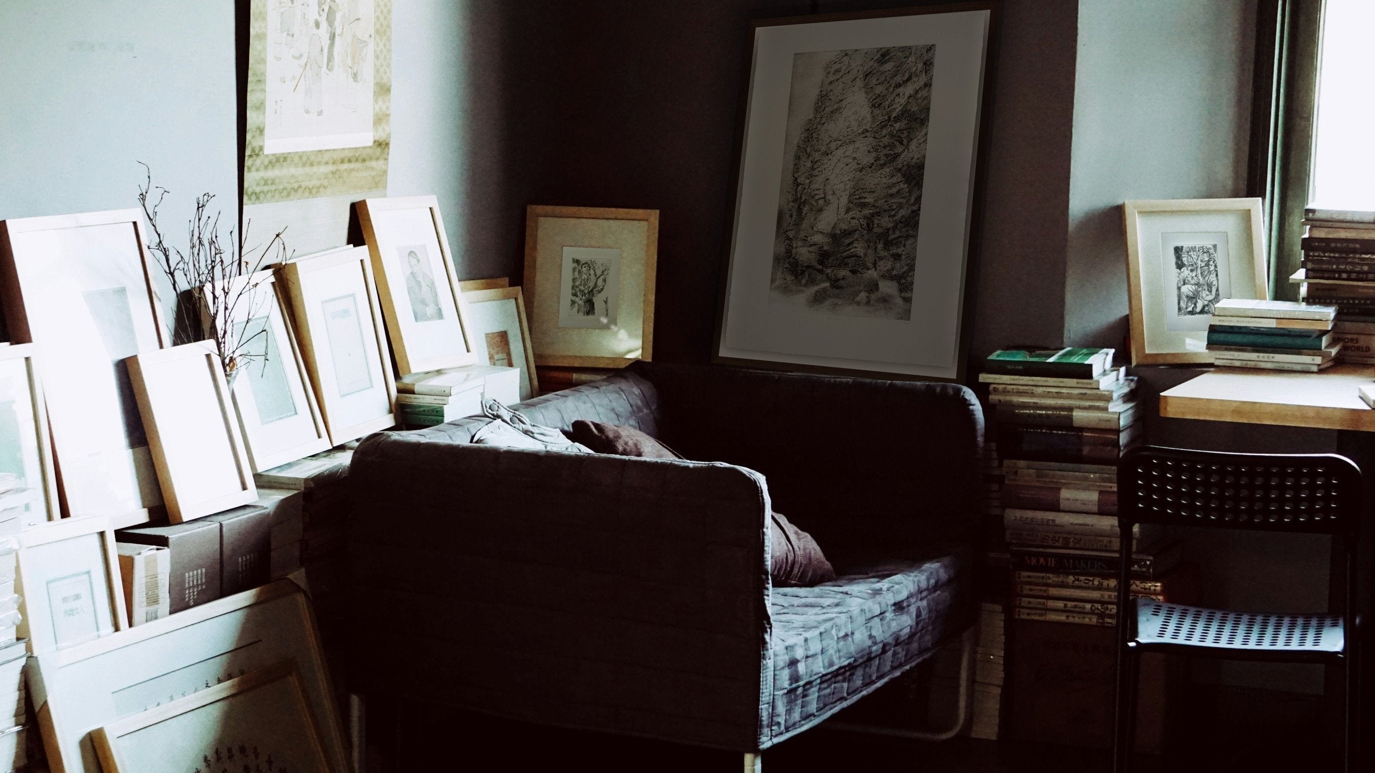 View of the interior of a room with a sofa, desk and chair. There are books on the desk and many framed artworks leaned against the walls of the room.