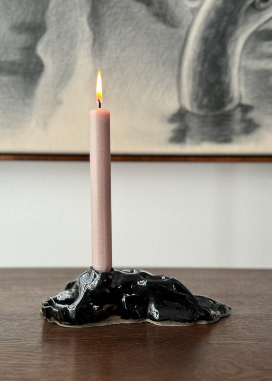 A black ceramic candle holder with a textured finish, resembling a geological formation, placed on a wooden surface with a blurred artwork in the background.