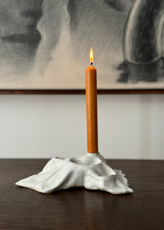 A ceramic candle holder with a white and glaze, designed by Aline Schwörer, displayed on a table with an orange candle lit inside.