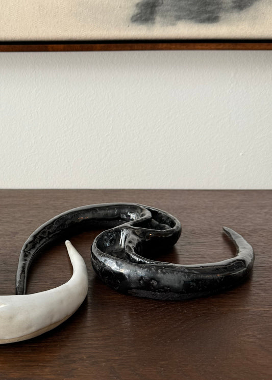 A unique ceramic sculpture of an indeterminate creature with a black glaze, displayed on a wooden surface with a blurred abstract painting in the background.