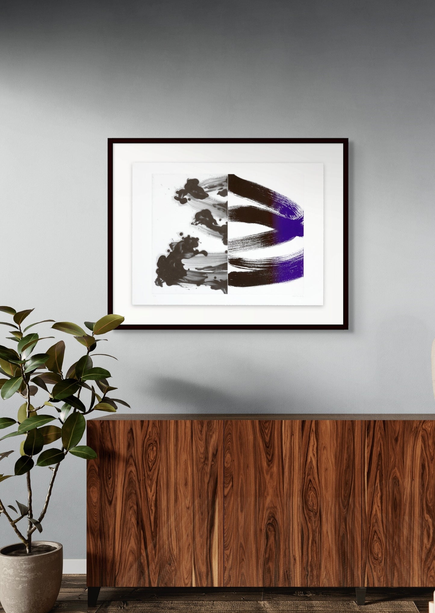 Framed abstract artwork on a gray wall above a wooden cabinet with a plant to the left.