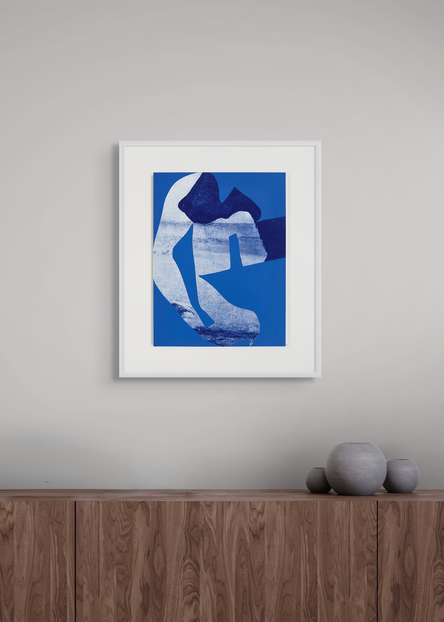 Framed abstract etching on a grey wall above a wooden cabinet with vases. 