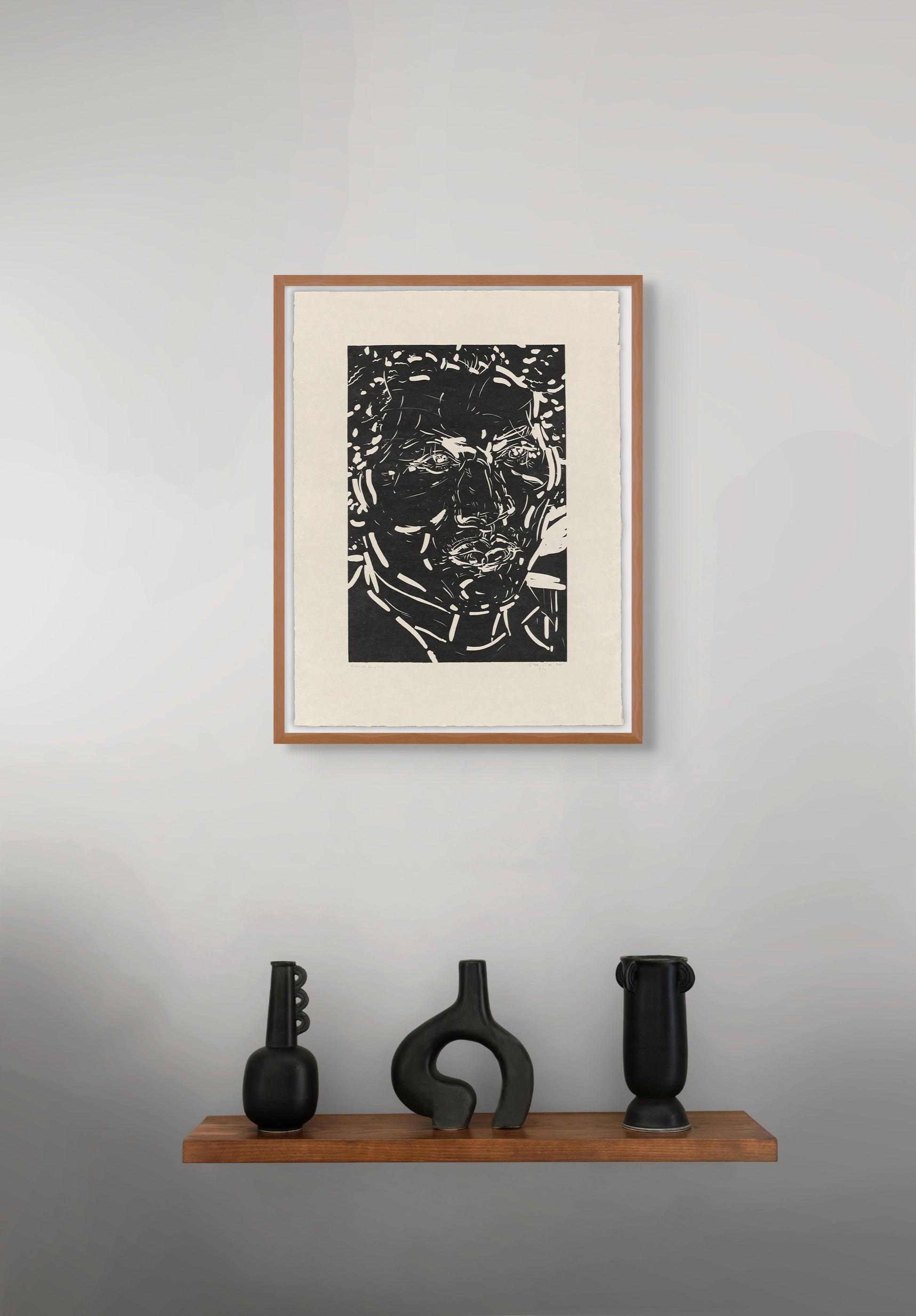 Framed black and white portrait by Elizabeth Peyton on a gray wall with three black sculptures on a wooden shelf below.