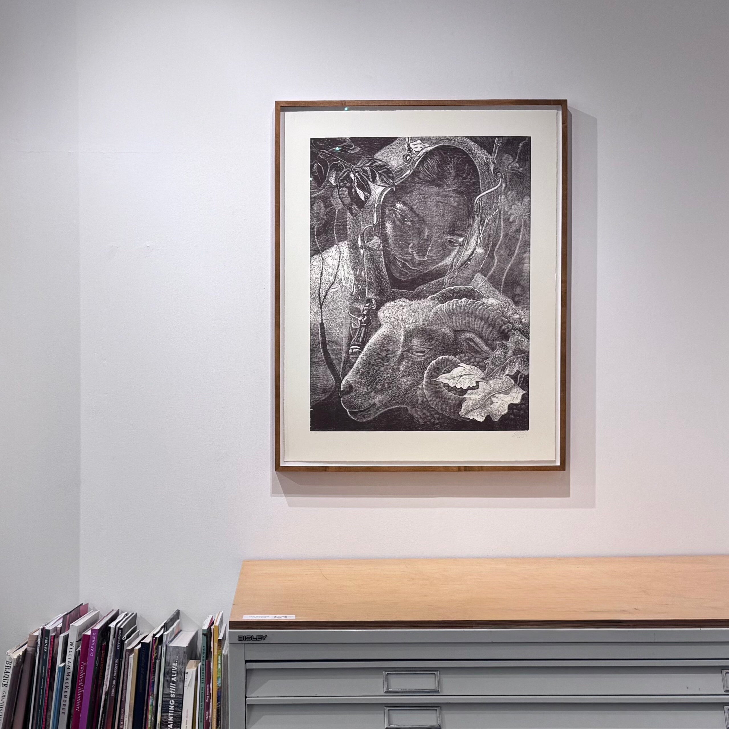 Framed black and white artwork by Dennis Scholl of a person in a helmet and sheep on a grey wall.