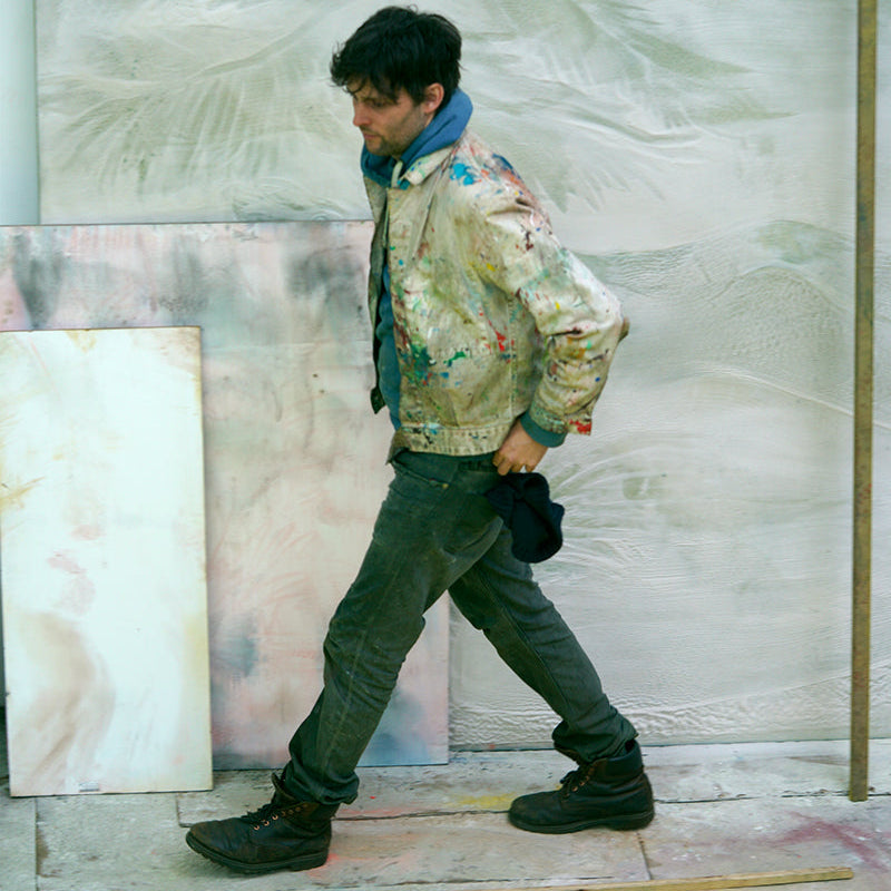 An artist walks across a studio floor wearing a heavily splattered cream-colored bomber jacket, dark pants, and a blue scarf, with large abstract canvases behind him .